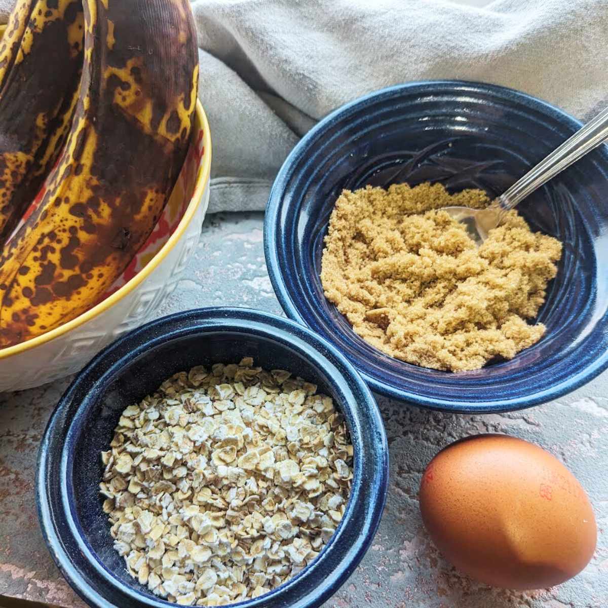 Individual bowls with ripe bananas, oats, brown sugar and an egg sit on a kitchen worktop.