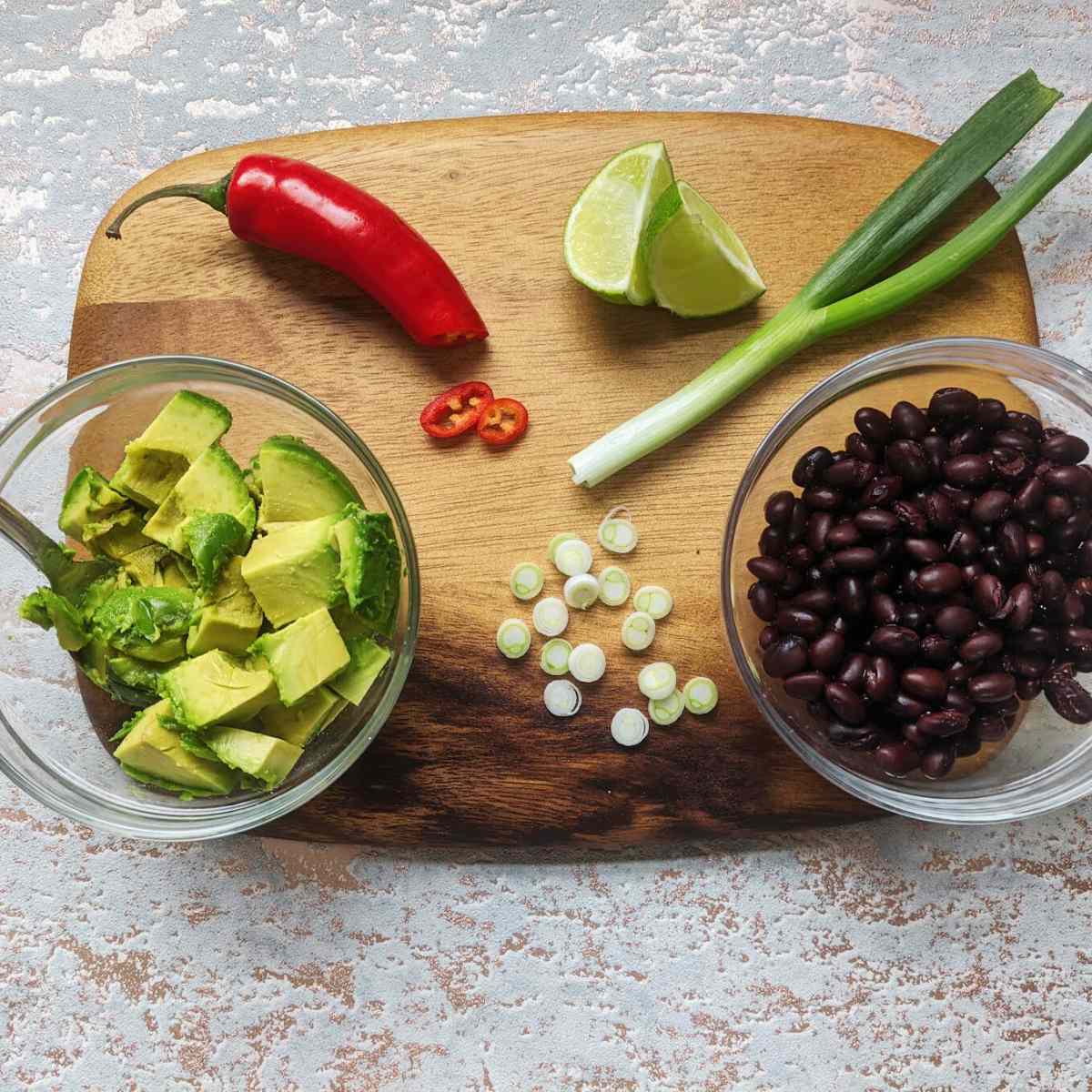 A chopping board with chopped avocado, spring onions, black beans, lime and chilli on top.