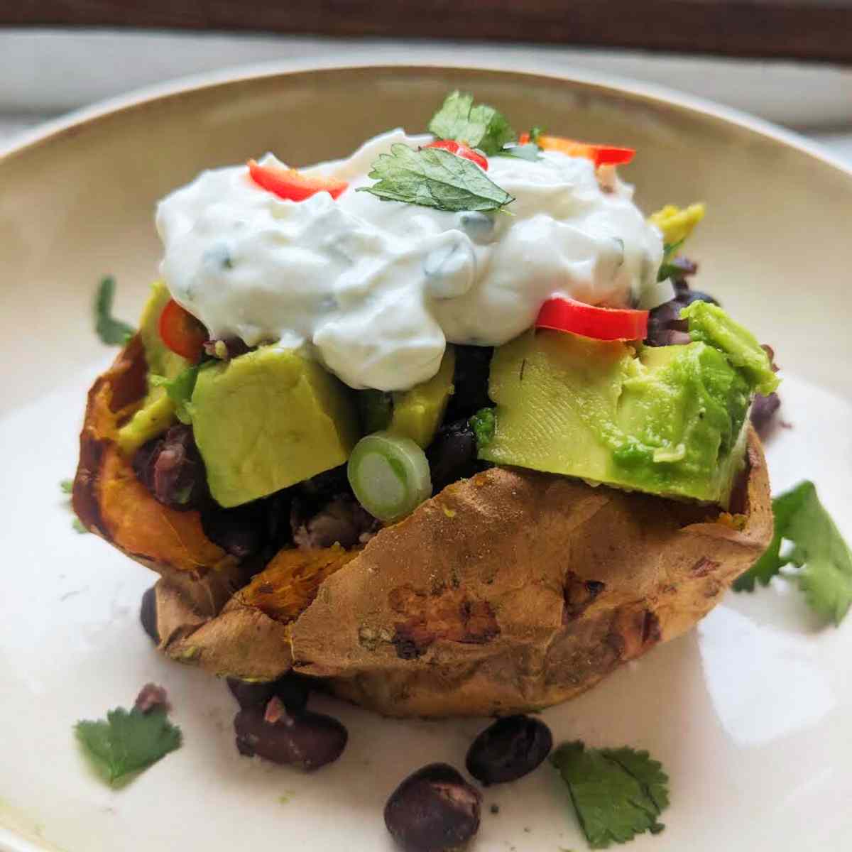 A baked whole sweet potato with avocado, black beans, spring onion, chilli, coriander and soured cream on top.