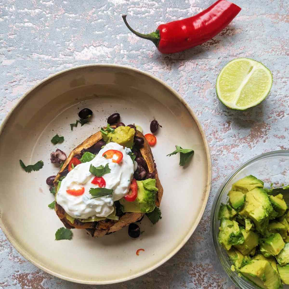 A cooked sweet potato in a bowl topped with black beans, avocado, red chili and coriander with a cut chili, lime and avocado at the side.
