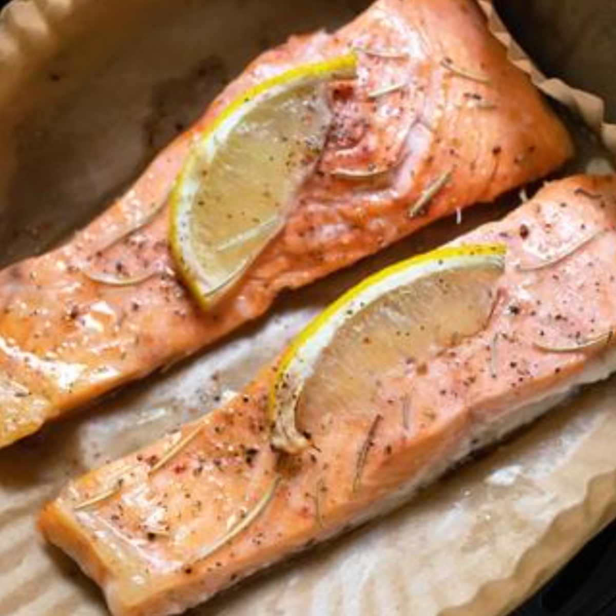 Two pieces of cooked salmon with black pepper and a wedge of lemon on each, in air fryer parchment