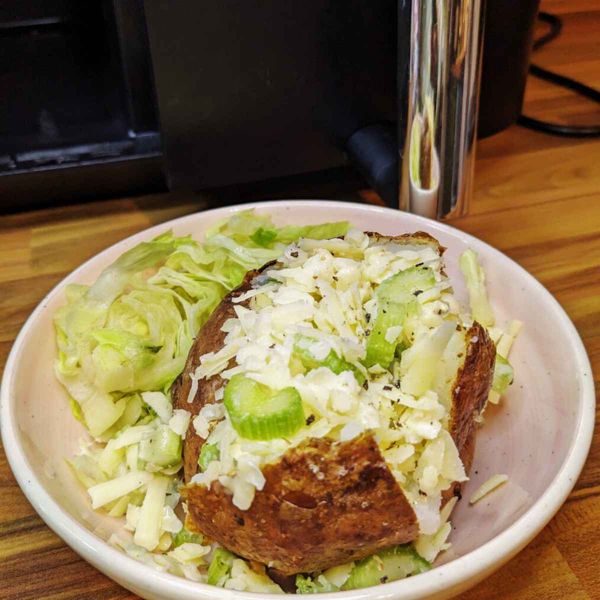 A baked potato sits in a bowl next to air fryer, with a topping of grated cheese and celery and shredded lettuce at the side..