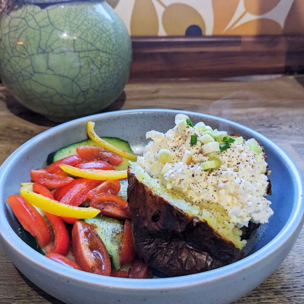 A baked potato topped with cottage cheese and spring onions sits in a bowl next to a chopped salad of cucumber, tomatoes and pepper.