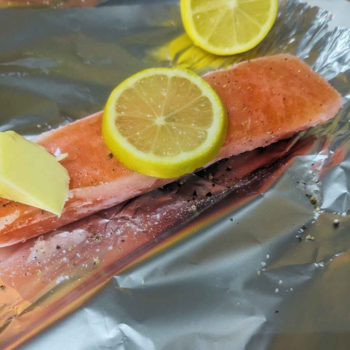 A frozen salmon fillet on a piece of aluminium foil with a slices of lemon and a pat of butter on top.