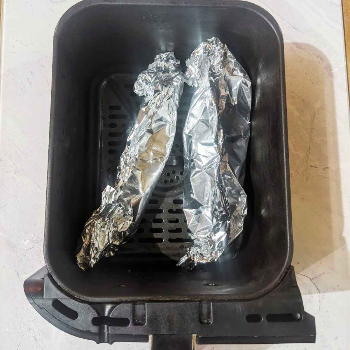 Two salmon fillets wrapped in aluminium foil inside an air fryer basket.