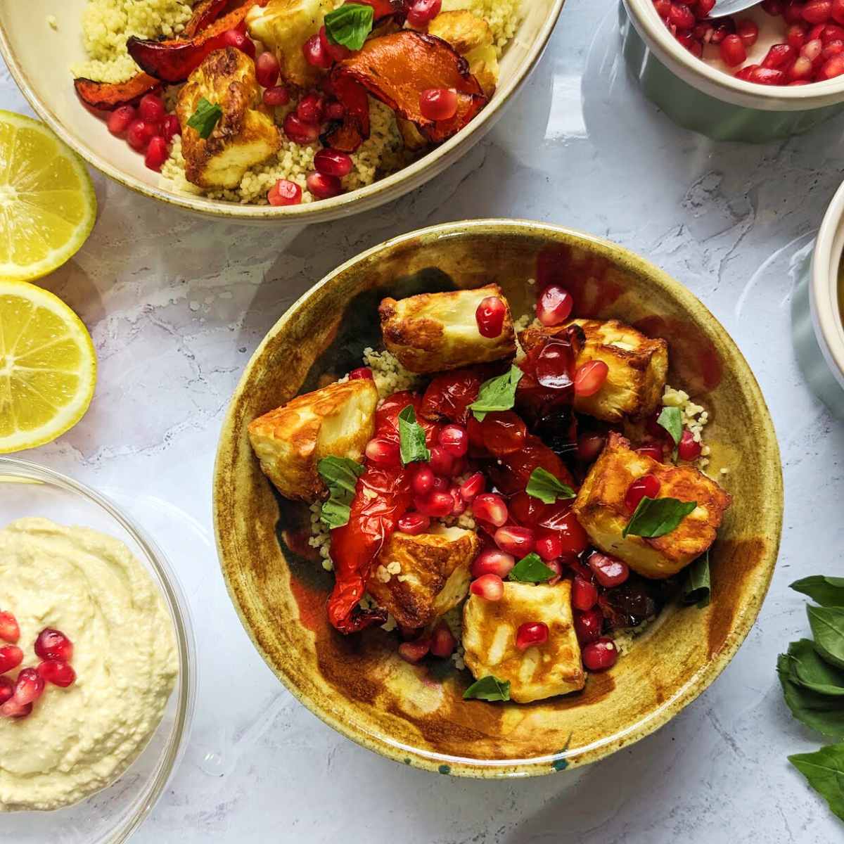 Couscous and halloumi salad topped with roasted red peppers and pomegranate seeds on a worktop with hummus, sliced lemons and herbs around the edge
