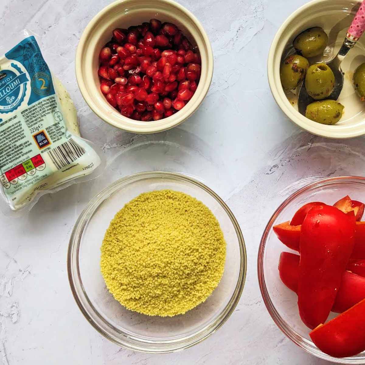 Ingredients for couscous and halloumi with a package of halloumi cheese, a bowl of dried couscous, a bowl of green olives, a bowl of sliced red bell peppers and a bowl of pomegranate seeds