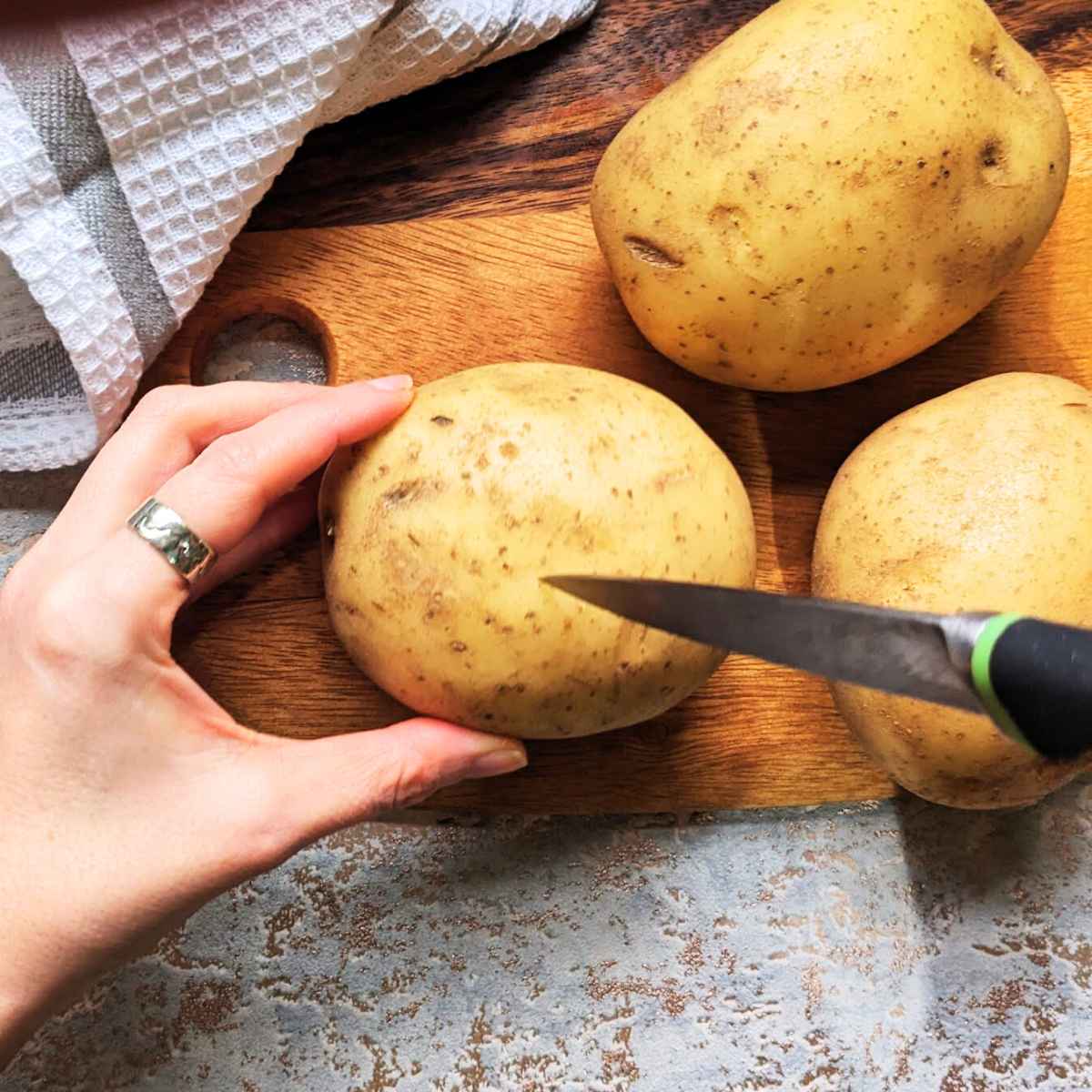 A hand holds a baking potato on a board while a knife is being used to prick the skin of the potato.