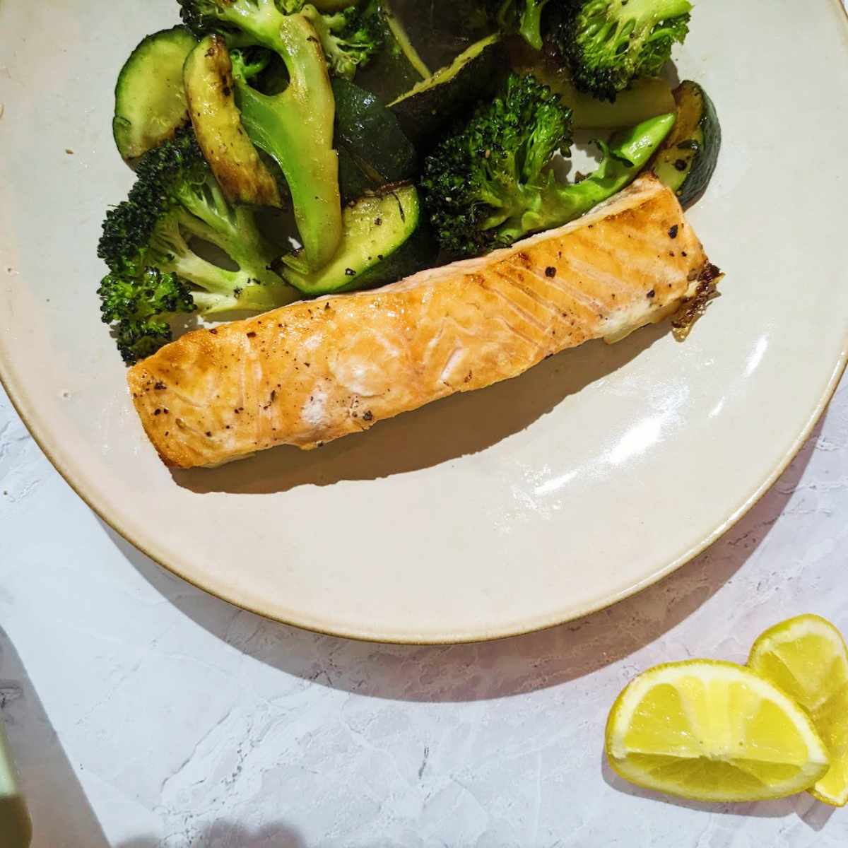 A salmon fillet on a white plate with green mixed vegetables and a wedge of lemon alongside.