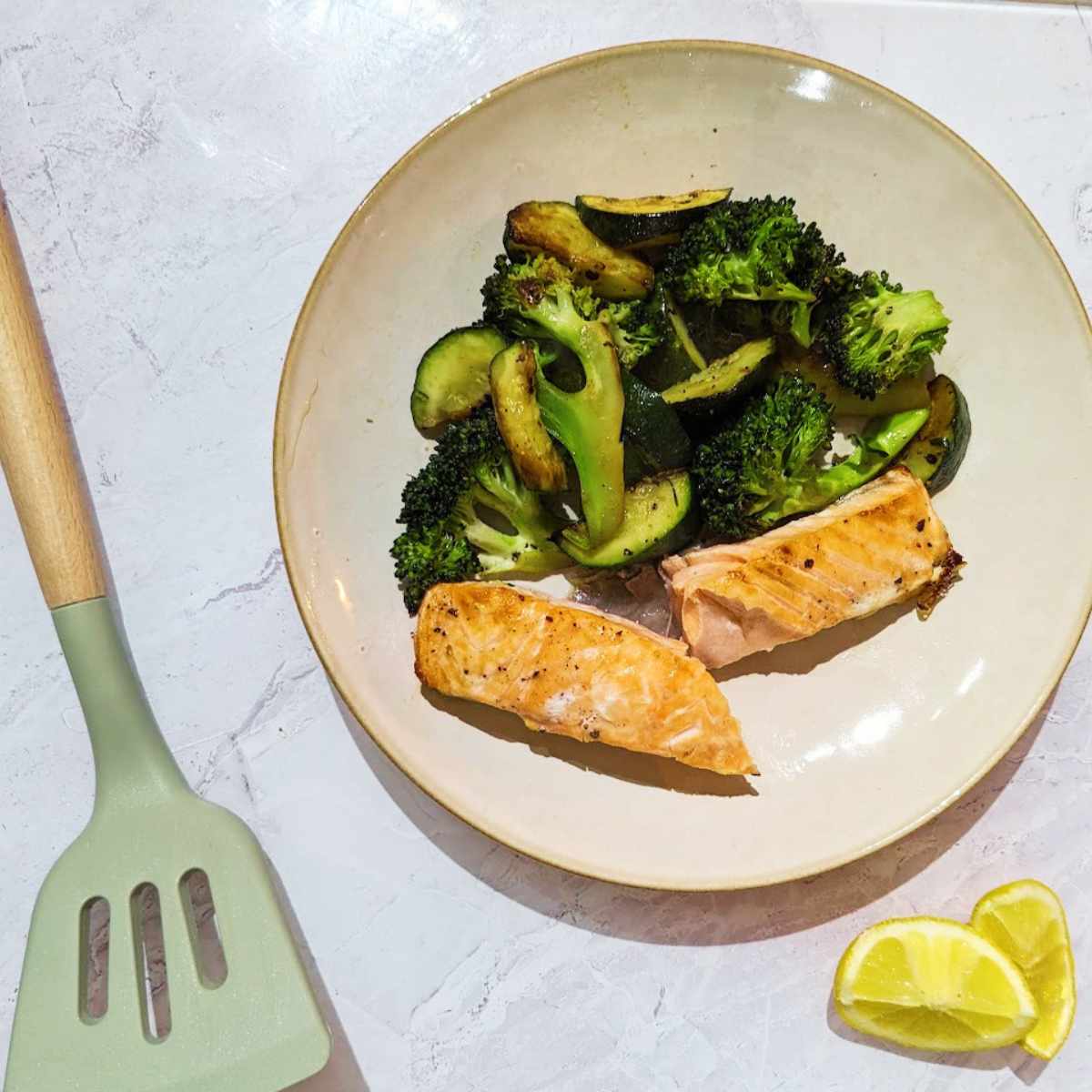 A salmon fillet on a plate with green mixed vegetables and a wedge of lemon alongside.