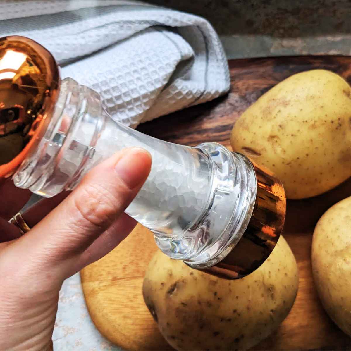 A hand holds a salt grinder over a baking potato which sits on a wooden board with two other potatoes.