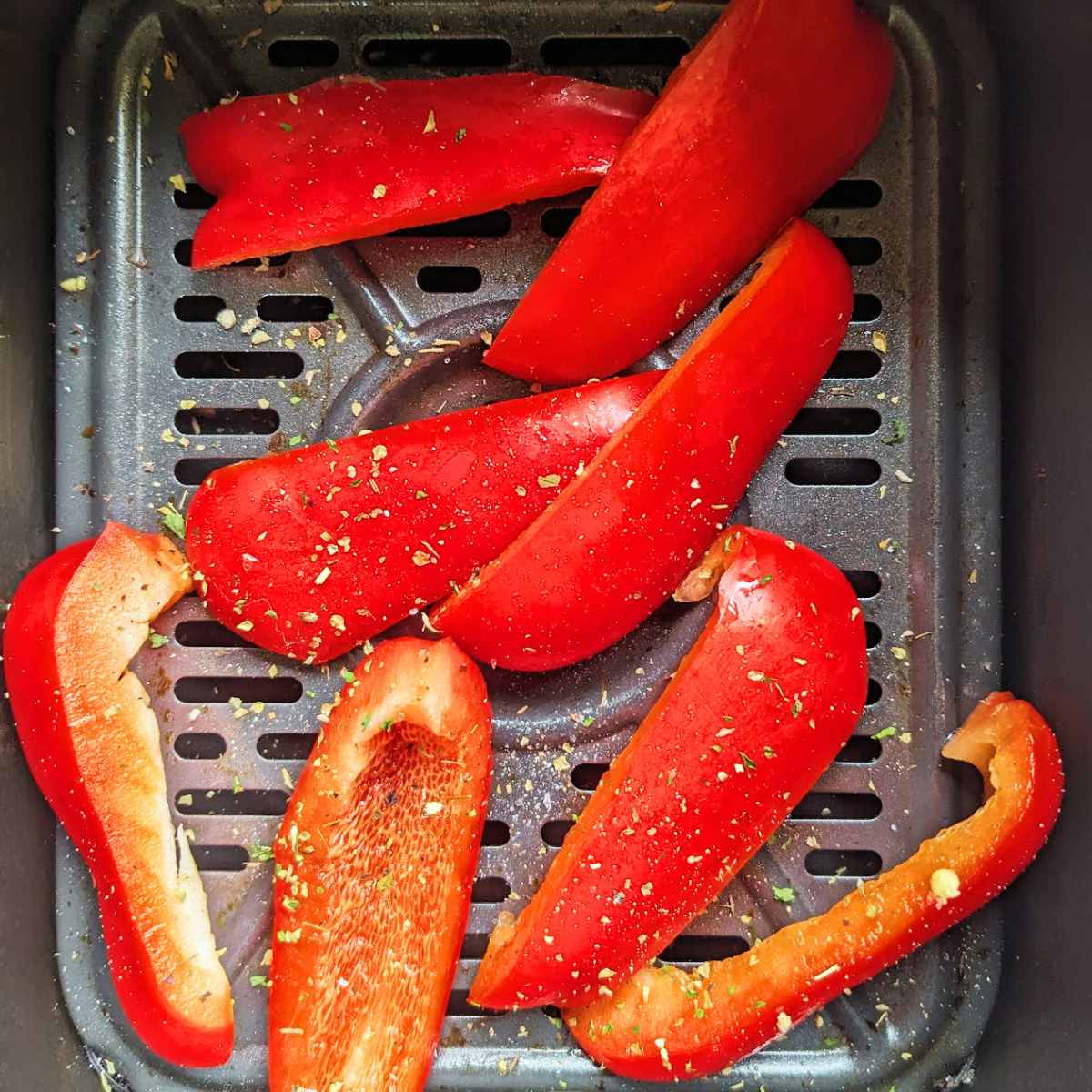 Sliced red bell peppers with dried herbs sit in an air fryer basket