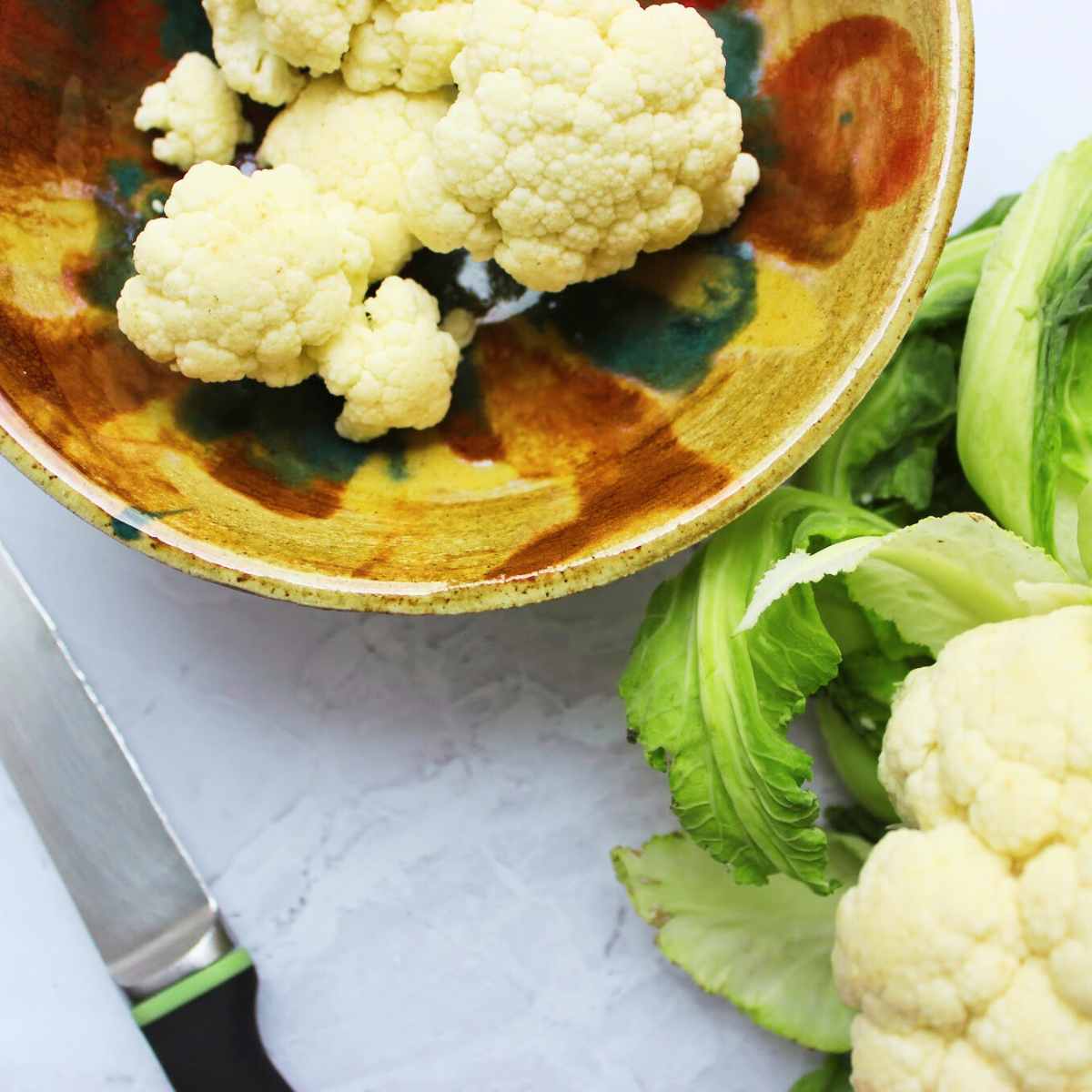 Cauliflower florets in a bowl with a head of cauliflower and a chef's knife by the side.