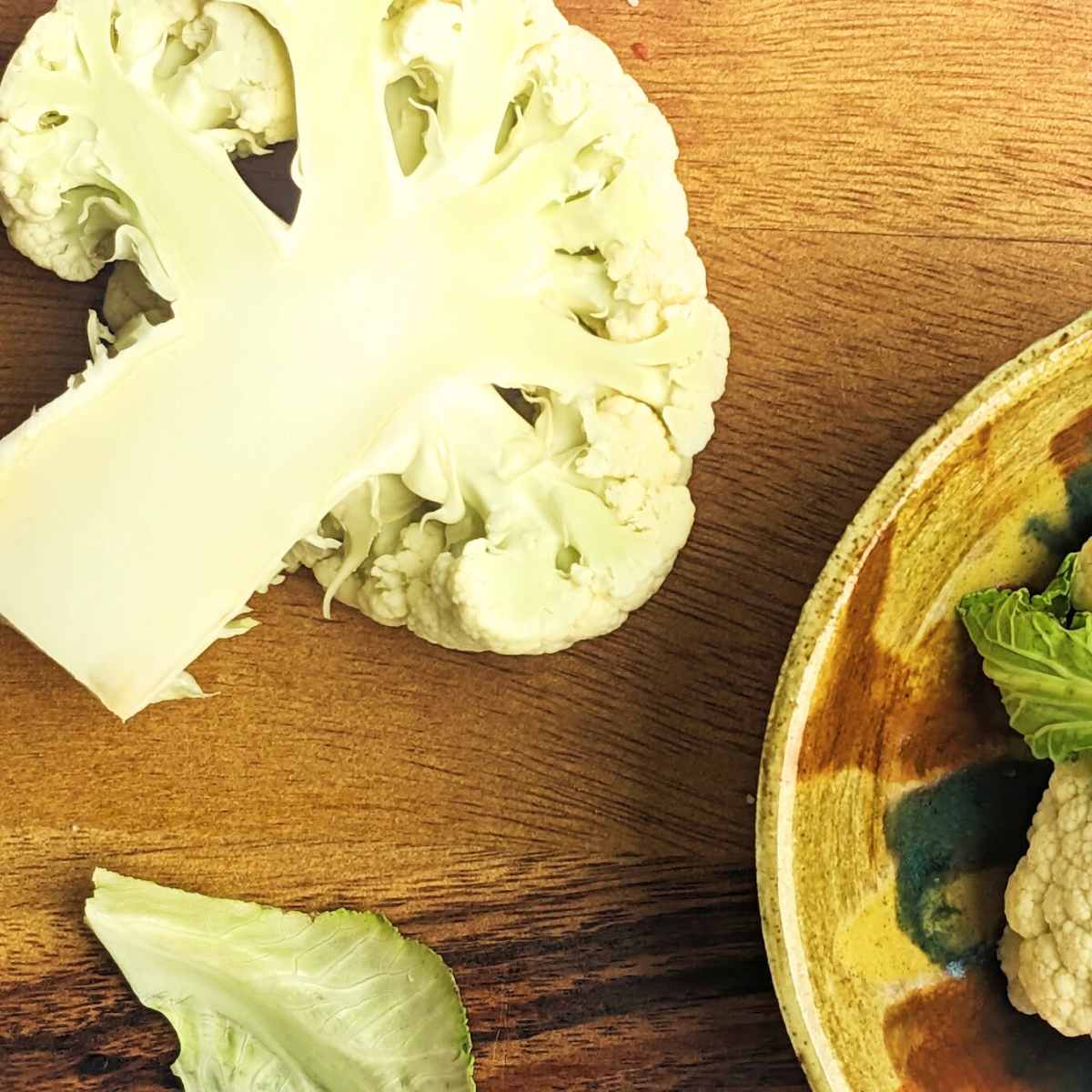 A slice of cauliflower on a chopping board with a bowl of cauliflower florets by the side.