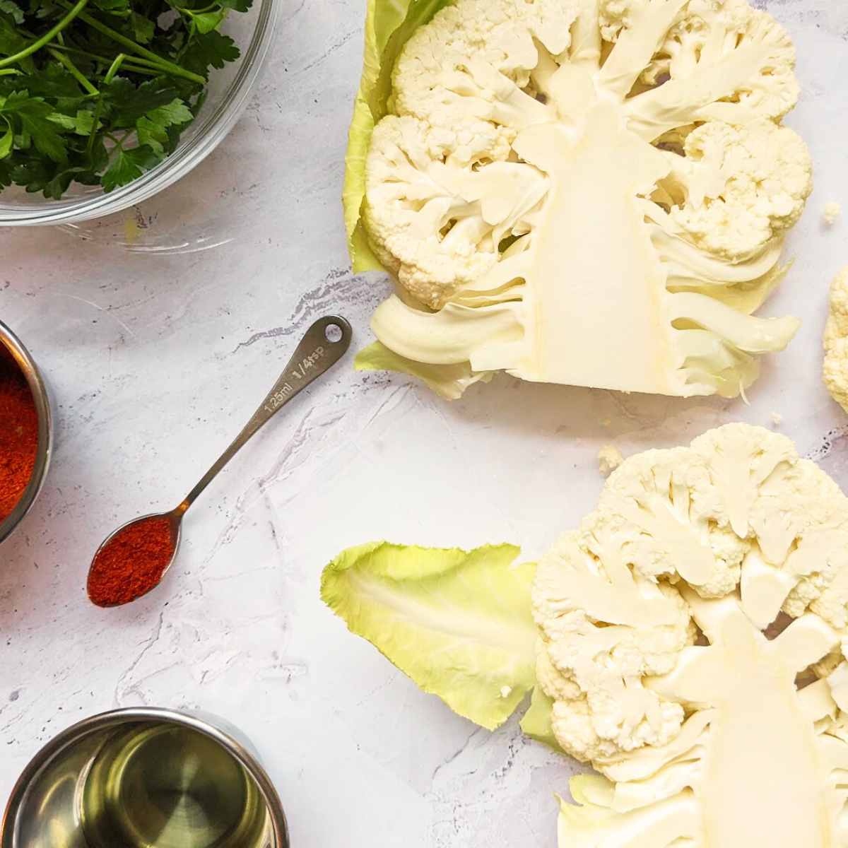 Cauliflower cut into steaks on a worktop with a pot of oil, a bowl of parsley and a teaspoon of paprika by the side.