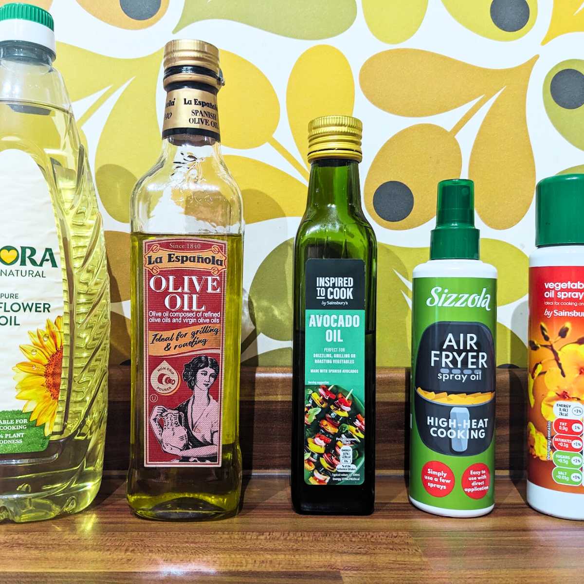 A selection of different cooking oils on a kitchen worktop. Selection includes sunflower oil, olive oil, avocado oil plus an air fryer spray oil bottle and a vegetable oil spray bottle.