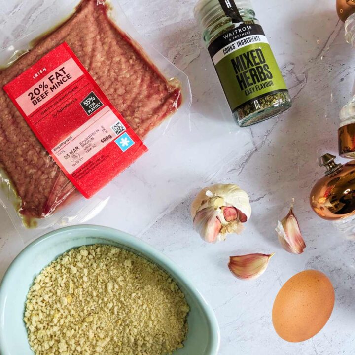 A white counter top with ingredients for meatballs on top. The ingredients include a pack of beef mince, white breadcrumbs, mixed herbs, garlic and an egg.