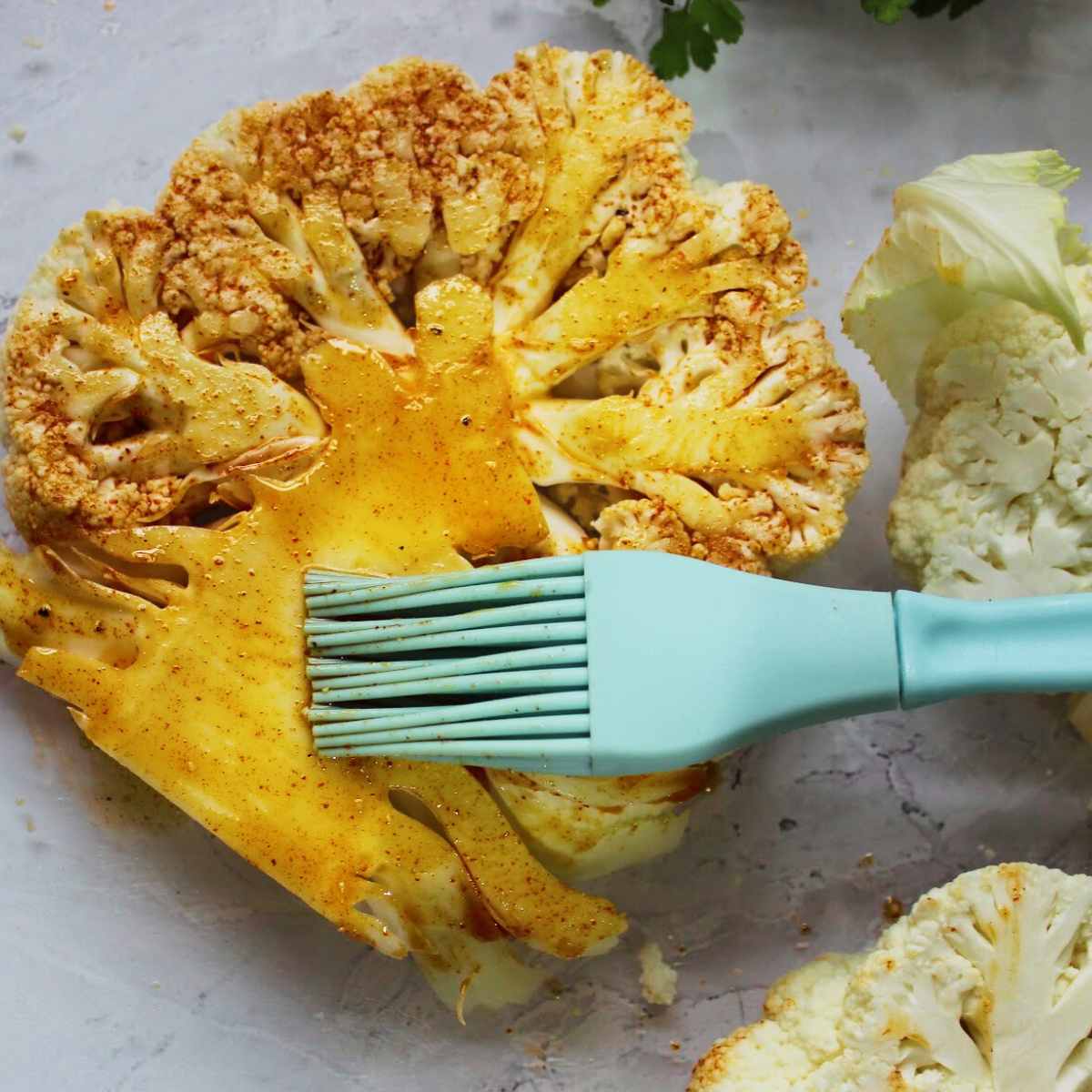 A pastry brush is being used to paint the side of a slice of cauliflower with an oil and paprika mixture.