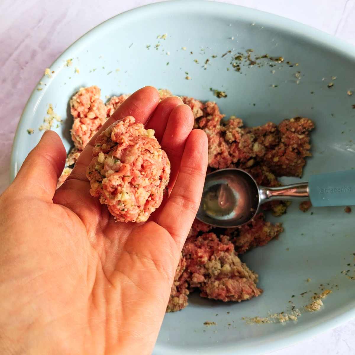 A hand is holding a freshly rolled meatball over a bowl that contains meatball mixture and an ice-cream scoop.