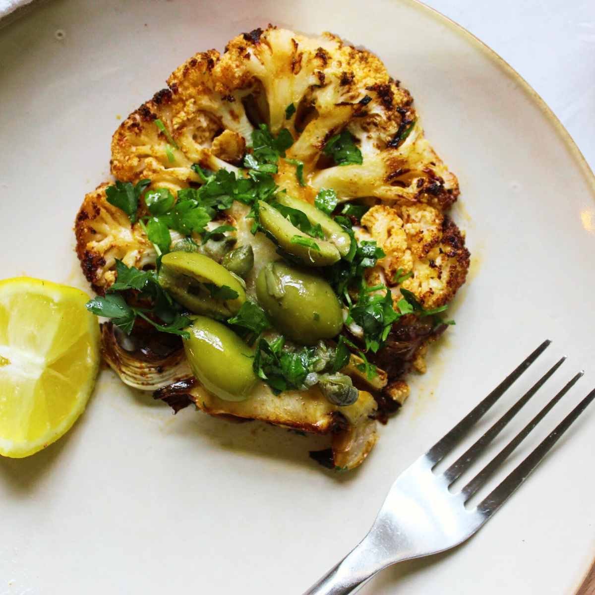 A cauliflower steak on a plate with olives, parsley and caper dressing.
