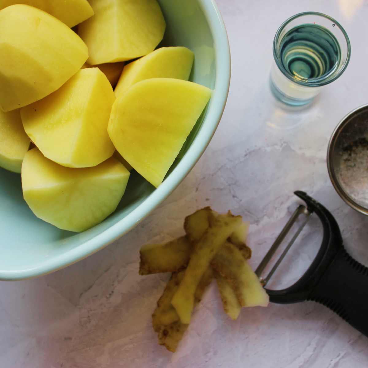 Peeled potato chunks in a bowl with a potato peeler, oil and seasoning alongside.
