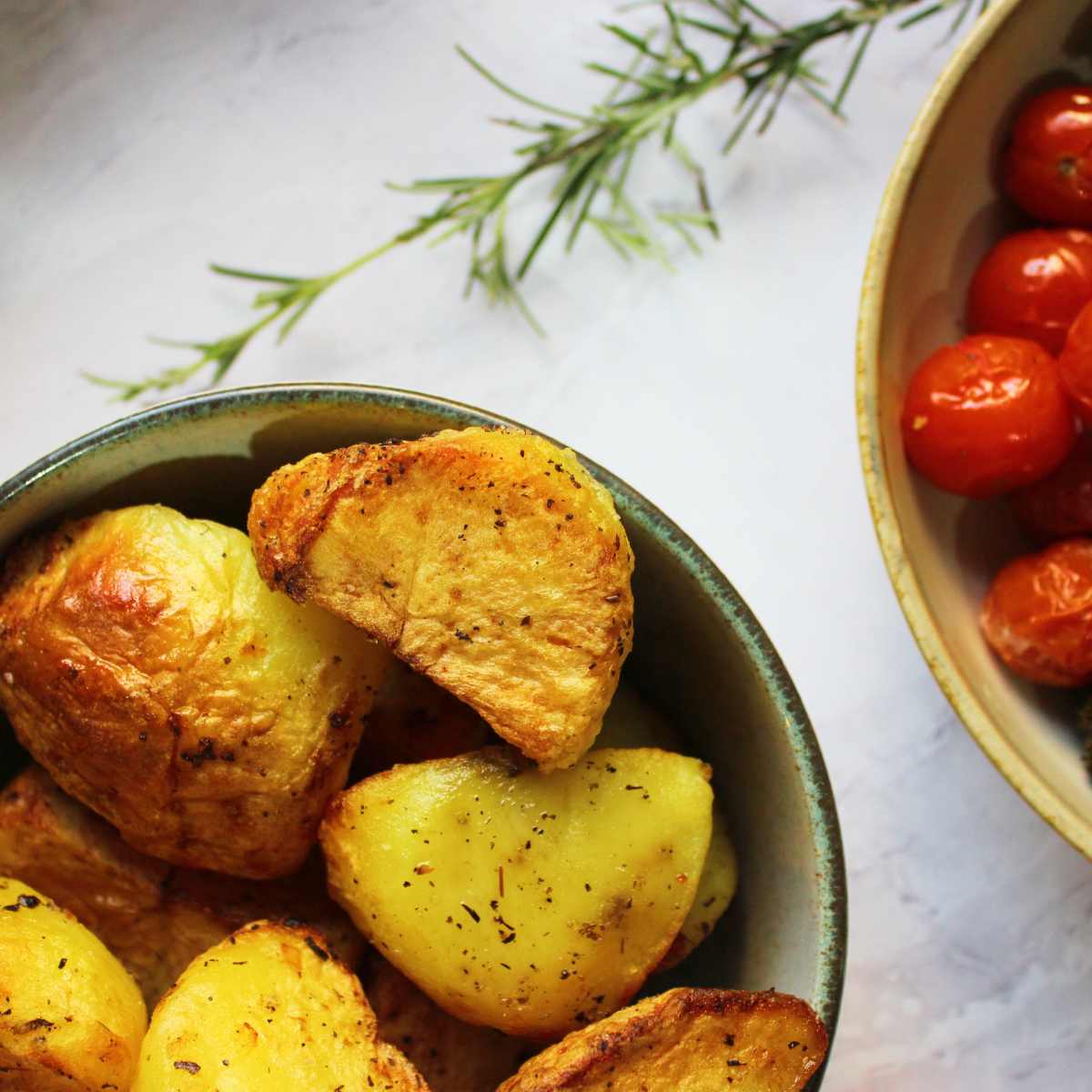 A bowl of air fryer roast potatoes with tomatoes and rosemary by the side.