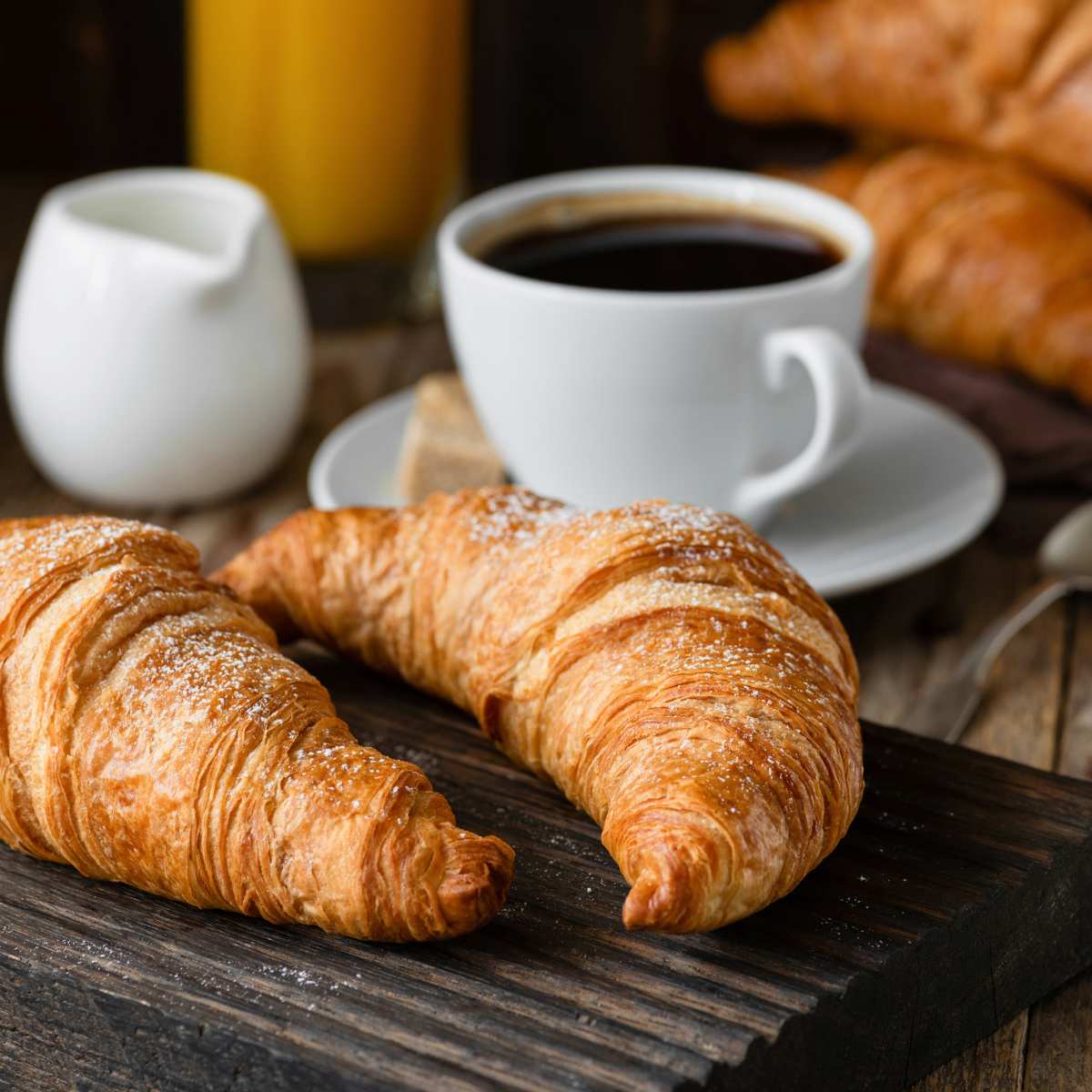 Two croissants on a board with a cup of black coffee and jug of cream in the background.