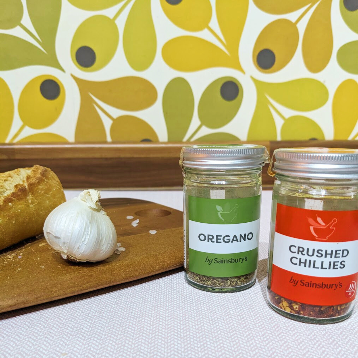 A jar of oregano and a jar of crushed chillies next to a chopping board with a baguette and a bulb of garlic.
