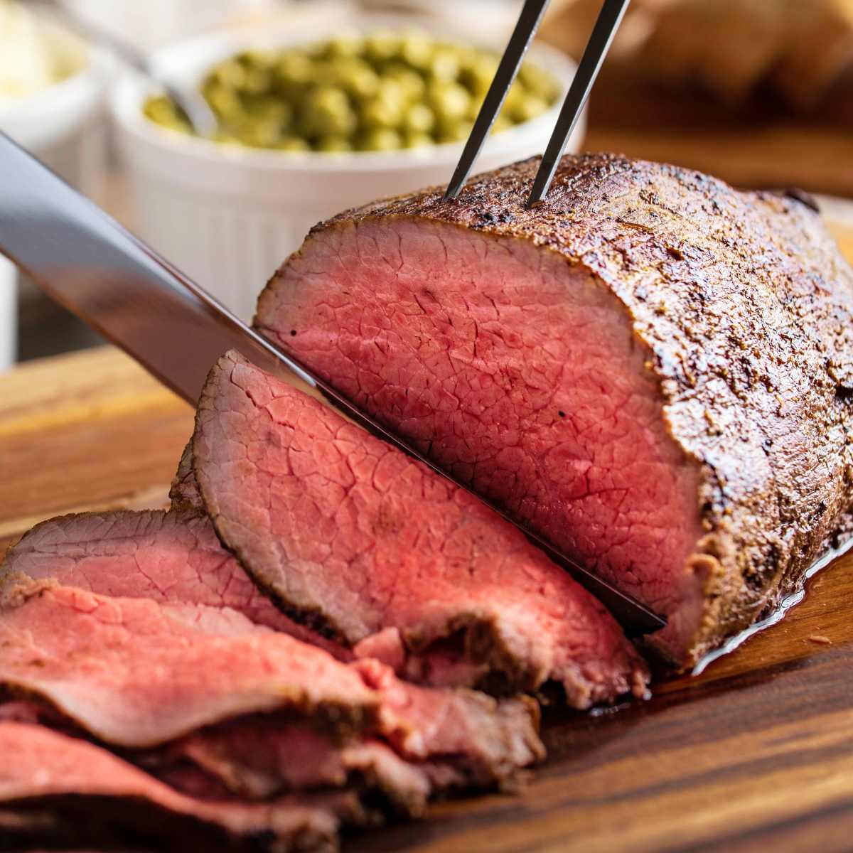 A roast beef joint being sliced on a wooden board with peas in a bowl in the background.