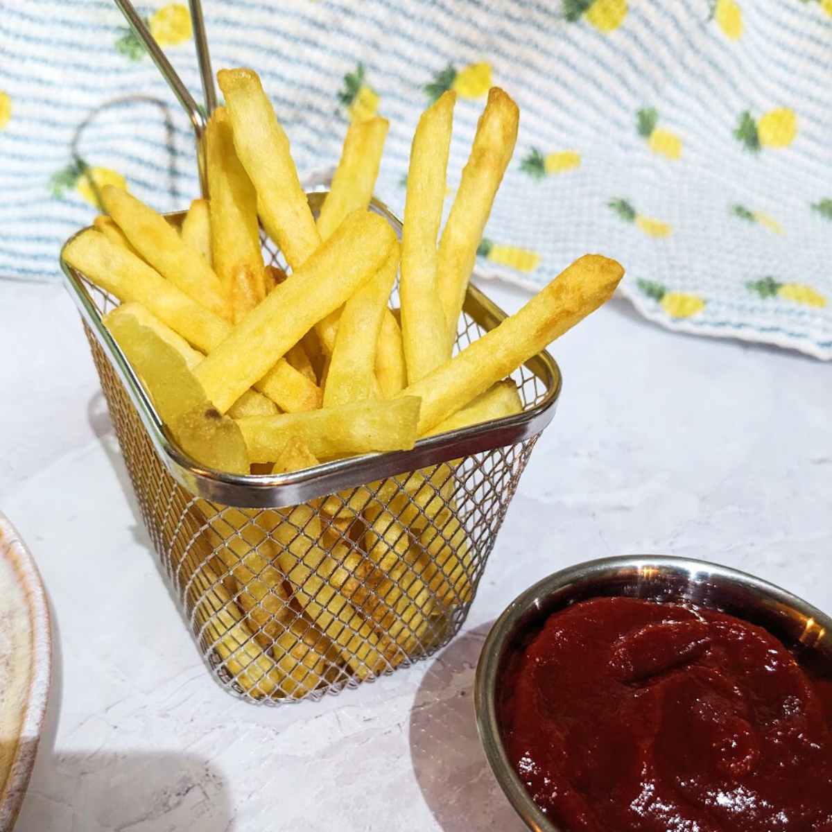 Cooked French Fries in a wire serving basket with a serving of mayo and ketchup at the side