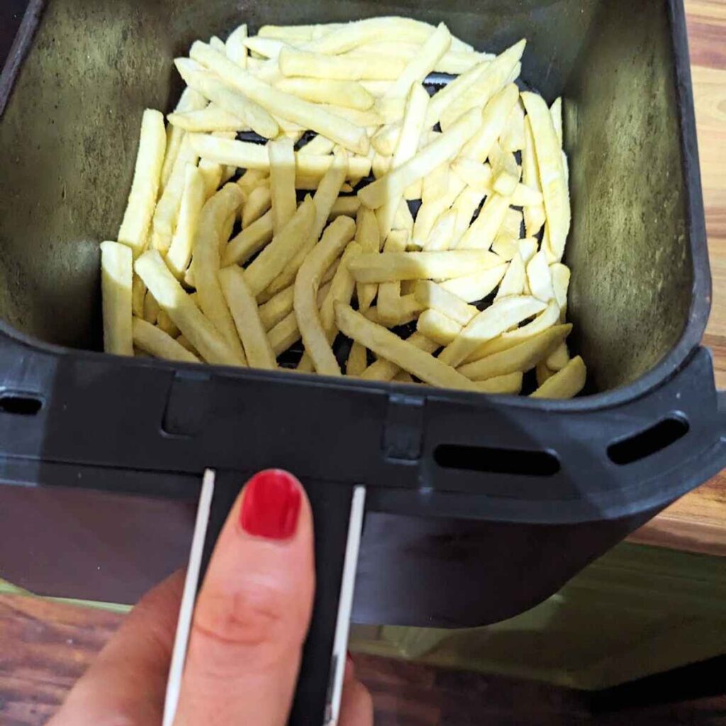 Frozen French fries in an air fryer basket with a hand on the handle.