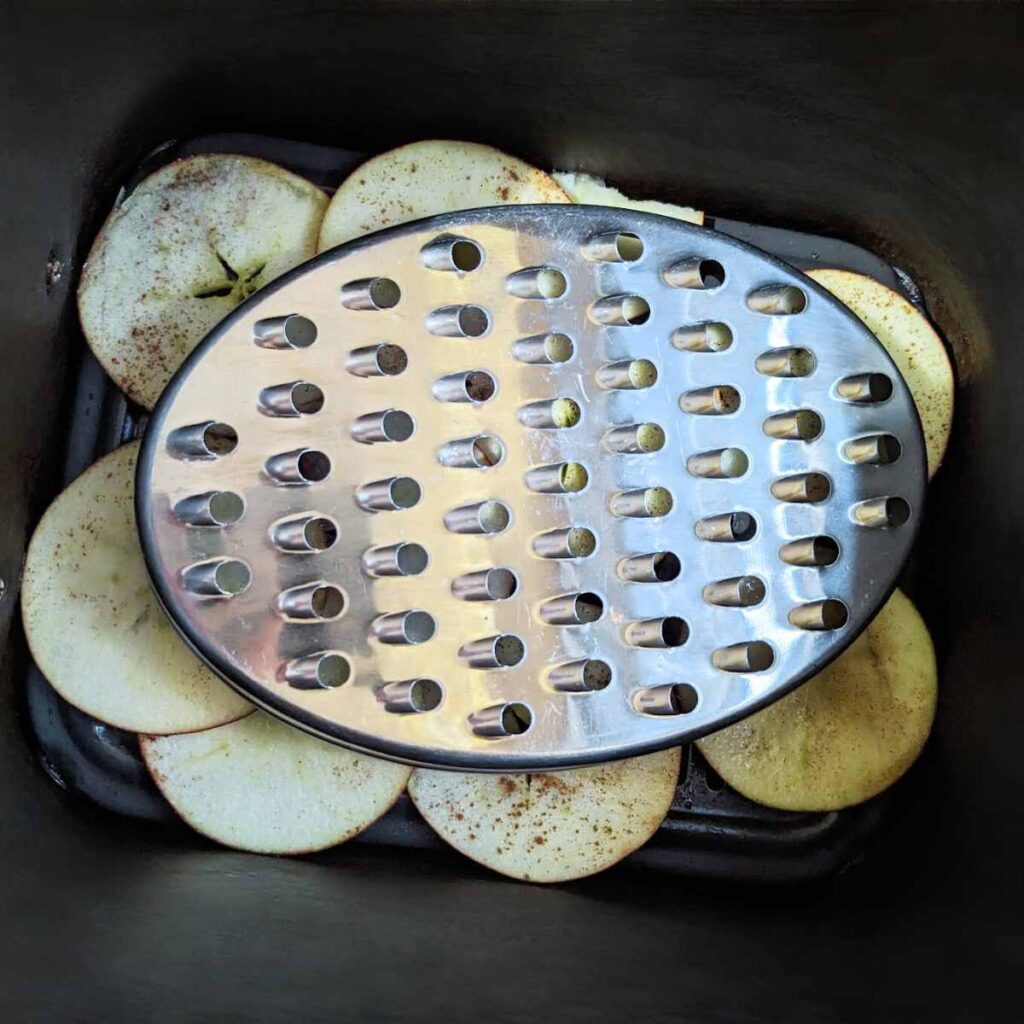 A metal cheese grater lid is used to weigh down apple slices in an air fryer basket.