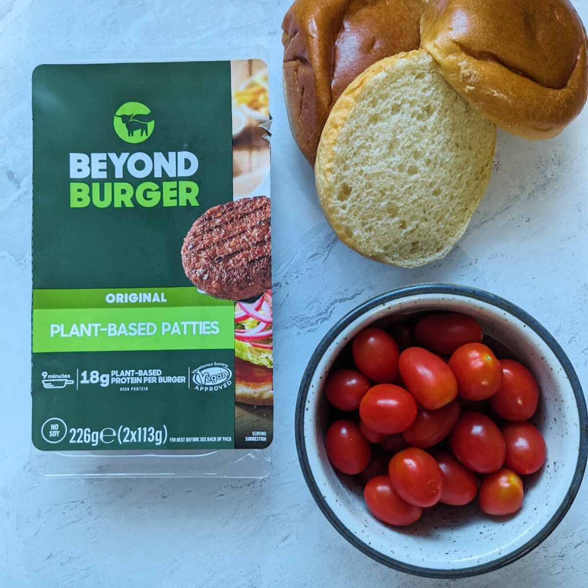 A pack of Beyond Burgers alongside sliced burger buns and a bowl of tomatoes