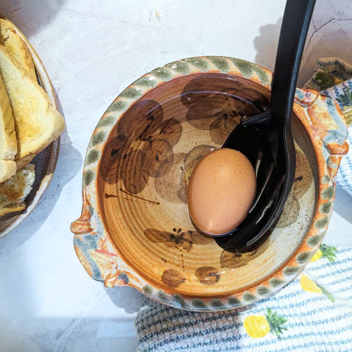 A boiled egg being lifted into a bowl of cold water using a silicone slotted spoon.