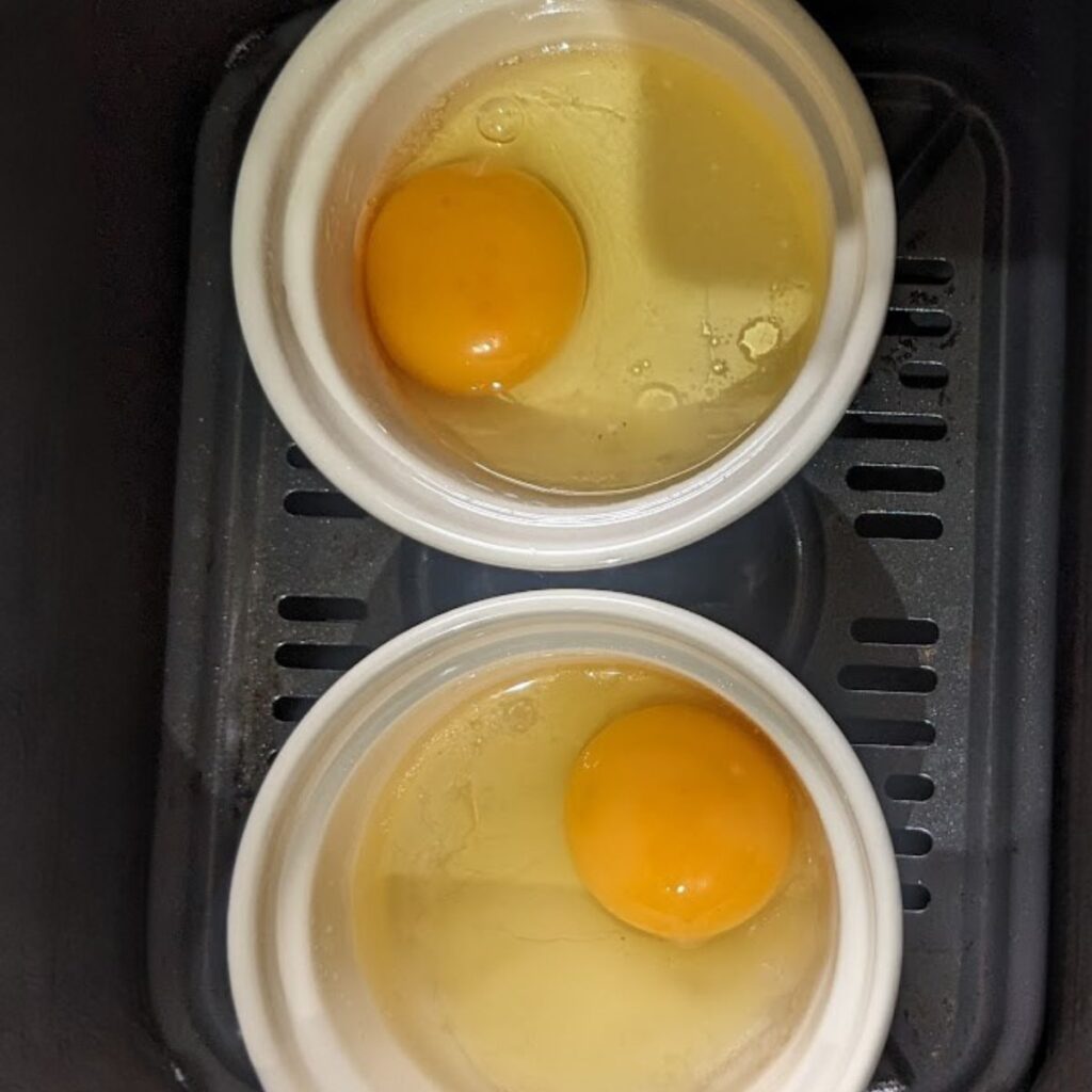 Two ramekins with a cracked egg in each inside an air fryer basket.