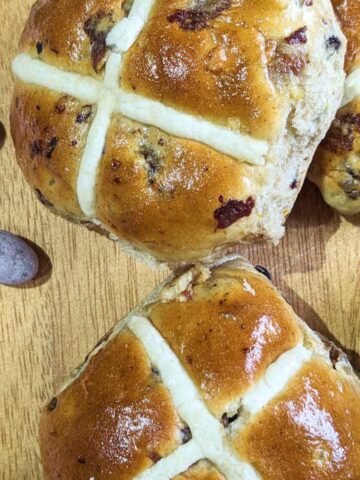 Two hot cross buns on a wooden chopping board with mini egg sweets at the side.