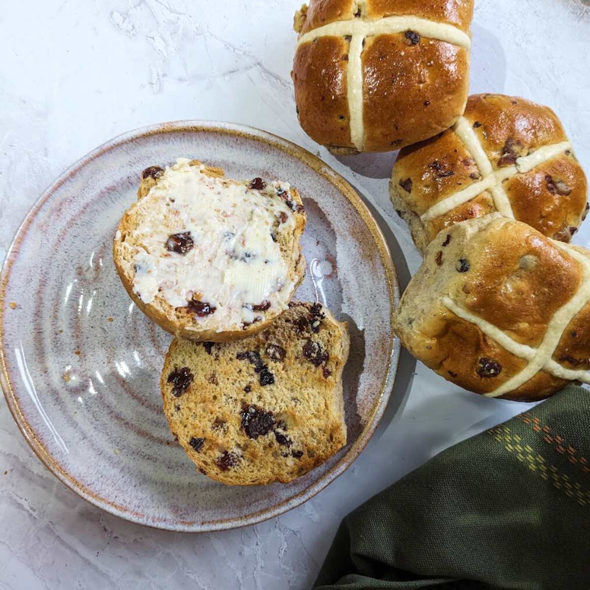 A buttered hot cross bun on a plate with a pile of three hot cross buns at the side.