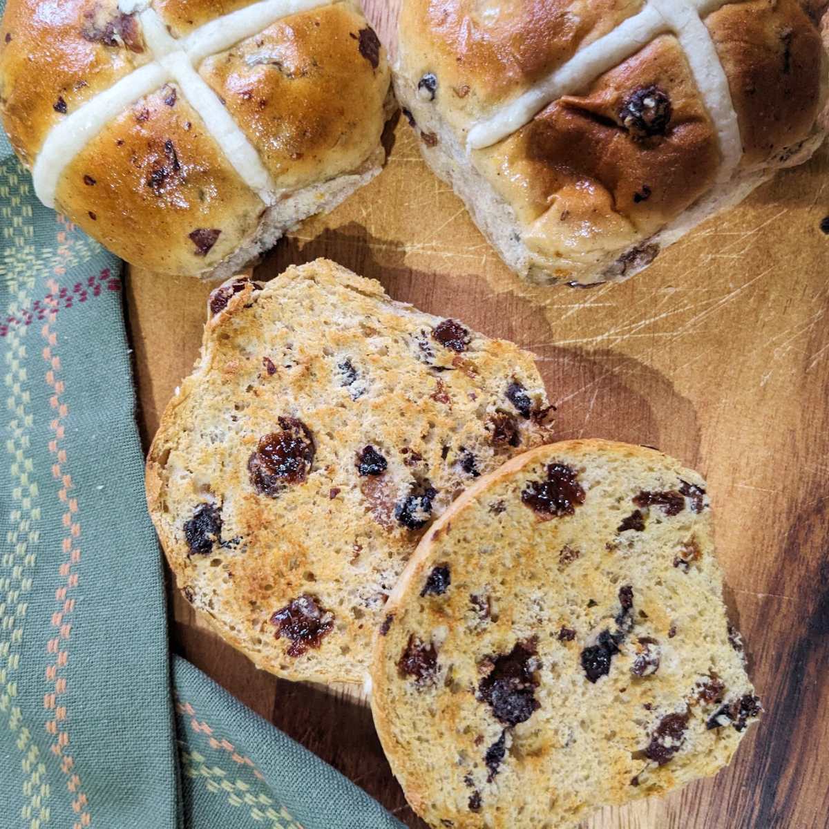 Hot cross buns on a chopping board with one bun sliced in half and toasted.