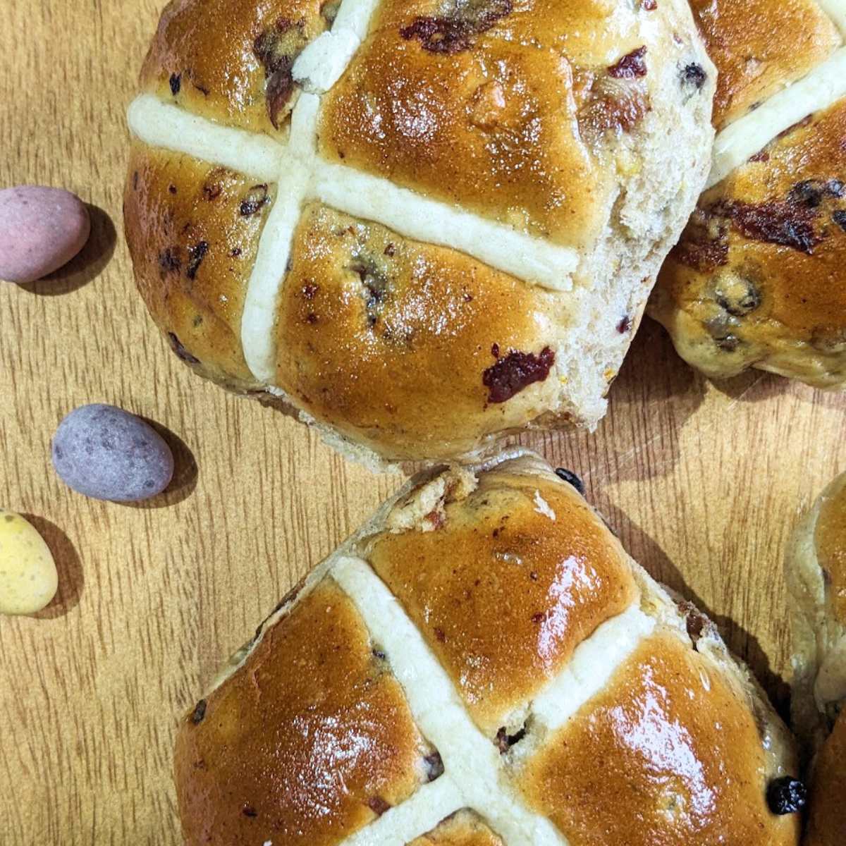 Two hot cross buns on a wooden chopping board with mini egg sweets at the side.
