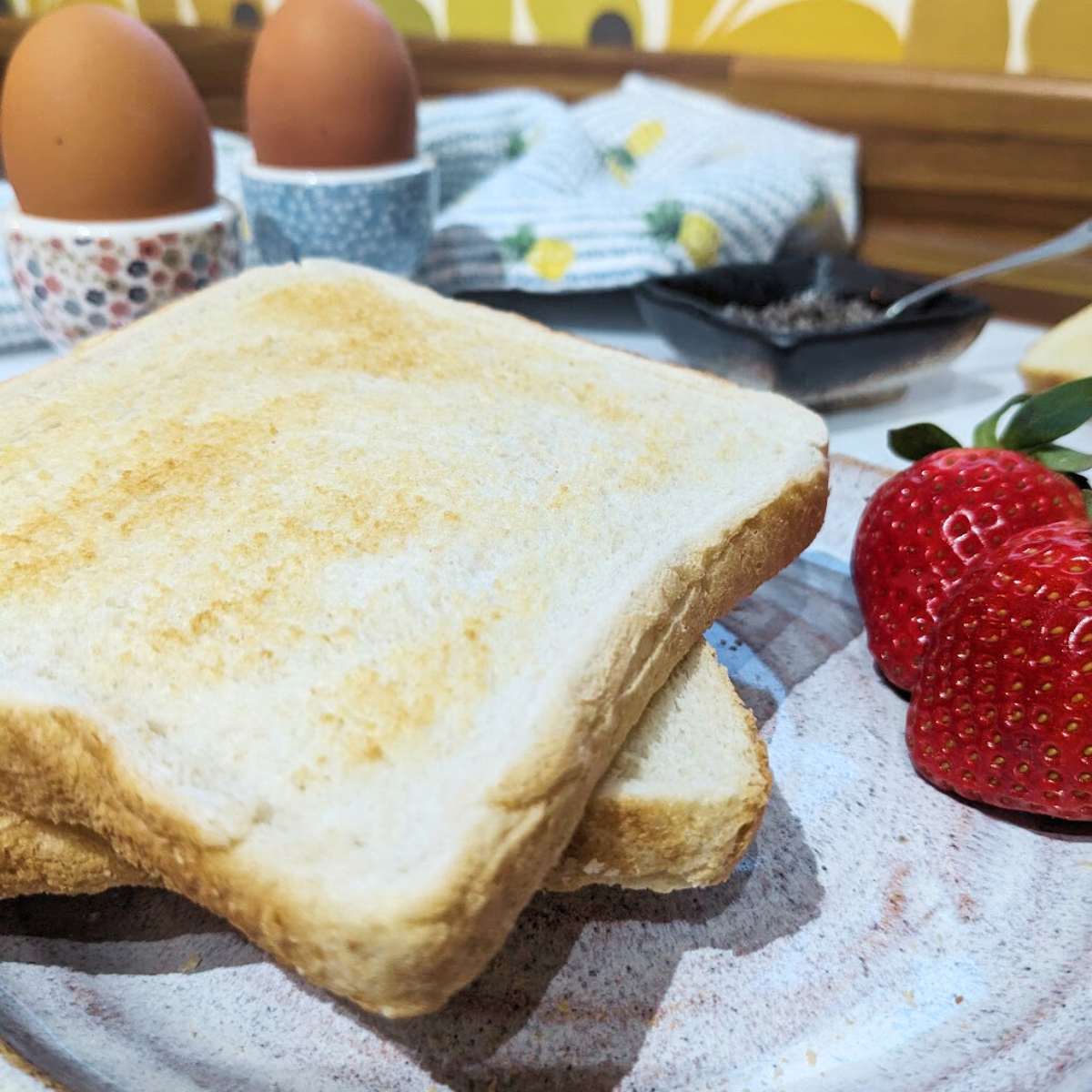 Two slices of toast on a plate. Two eggs in egg cups sit beside the plate. Two fresh strawberries are on the side of the plate.