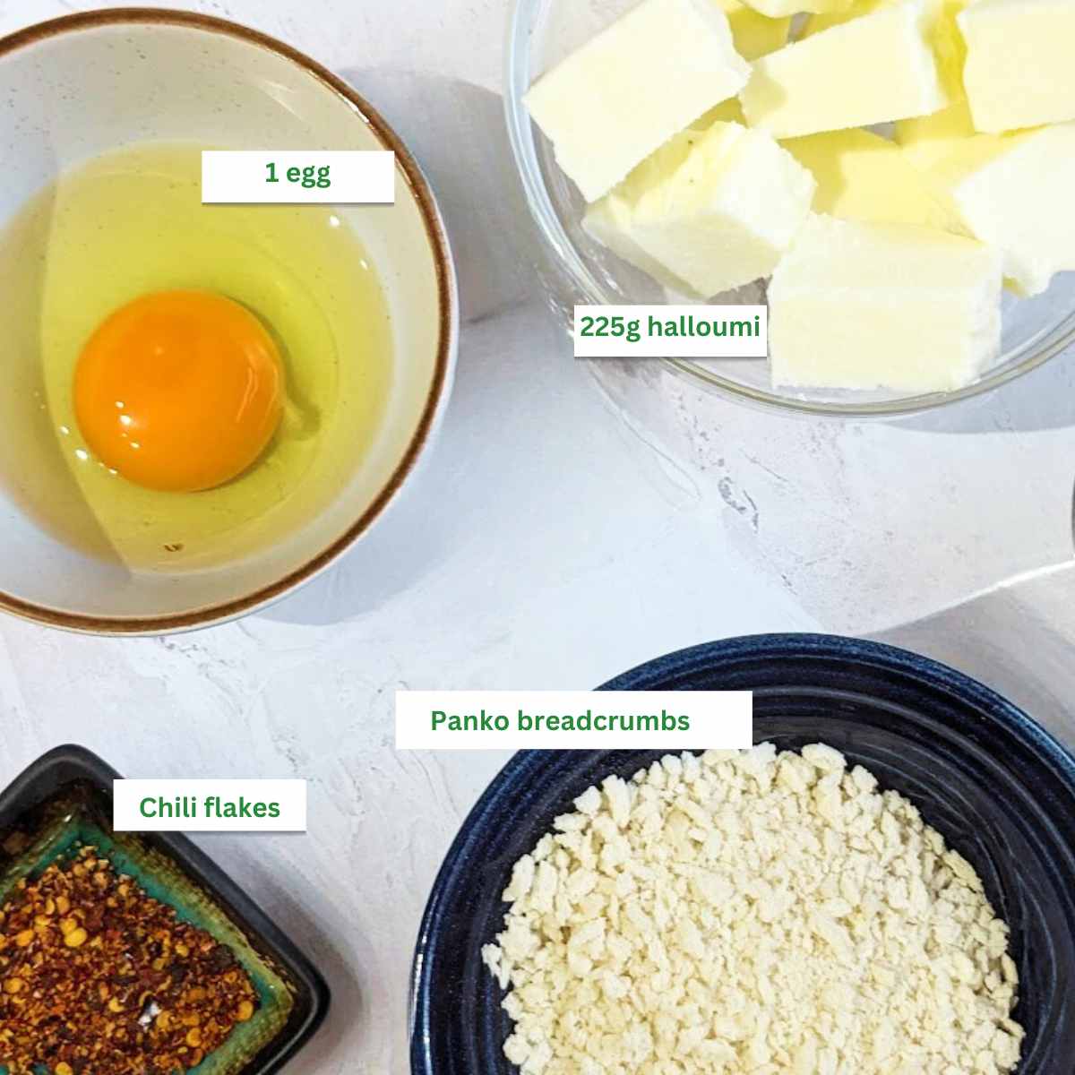 Ingredients for air fryer halloumi bites with chopped halloumi, an egg, chili flakes and panko breadcrumbs in individual bowls.