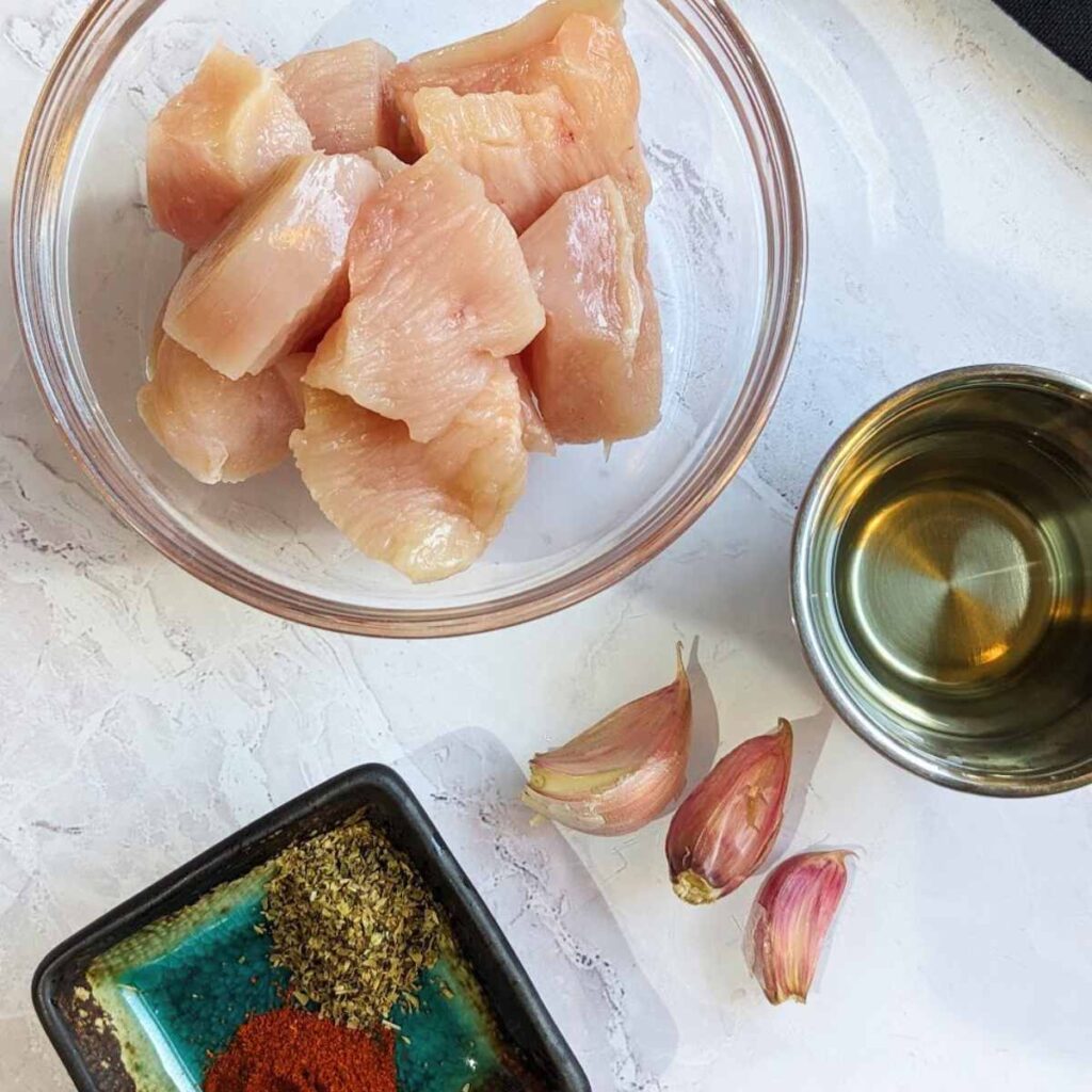 Ingredients for air fryer chicken bites including diced chicken breast in a glass bowl. Herbs, garlic and oil are laid out on the surface next to the bowl.