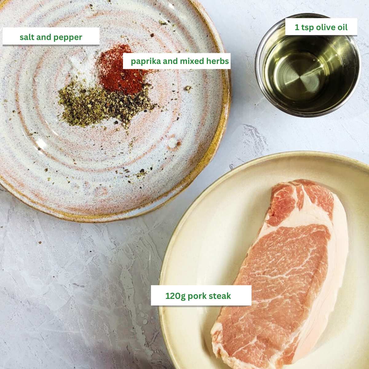 Ingredients for air fryer pork steak, includes a pork steak on a plate, olive oil in a small bowl and salt pepper, paprika and dried herbs on a plate.