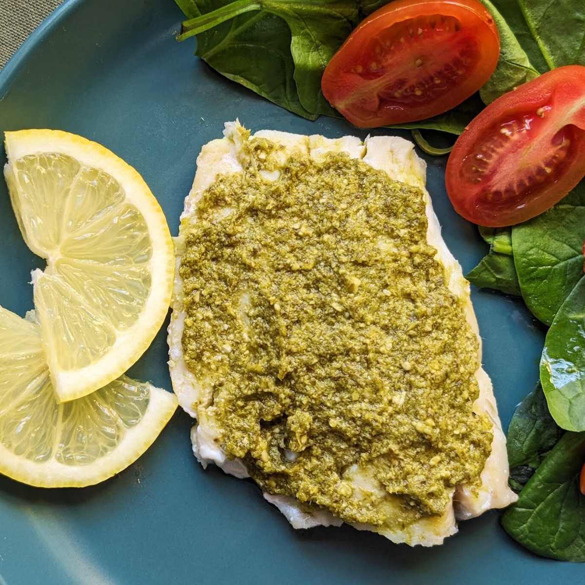 A haddock fillet with a pesto topping on a plate with a side salad of spinach and tomatoes.