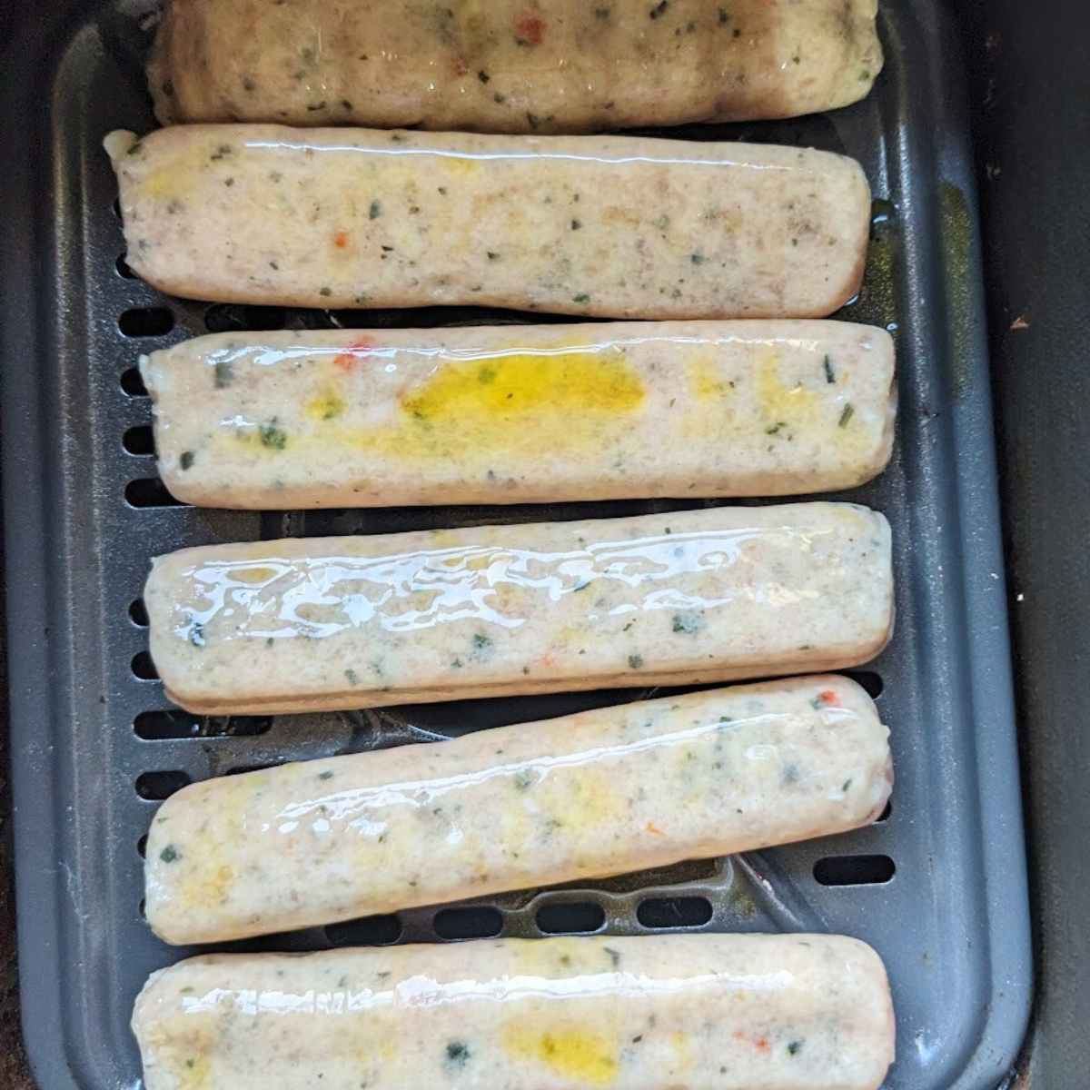 Uncooked chicken sausages lined up in an air fryer basket with a drizzle of oil on top.