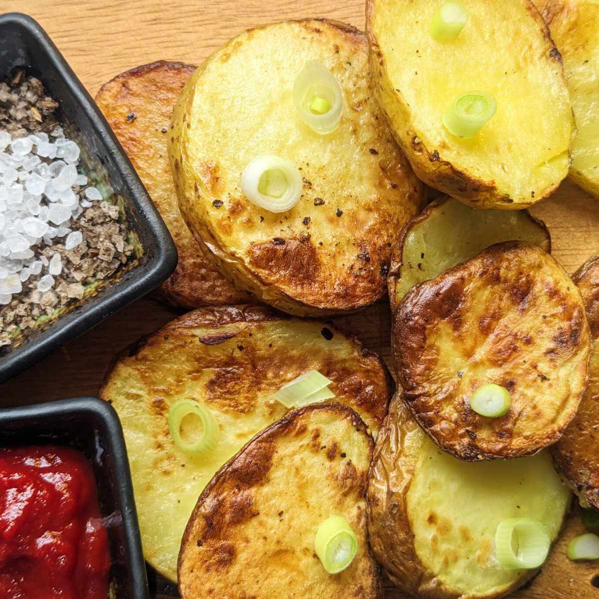Sliced potatoes on a board with seasoning and tomato sauce in bowls at the side.