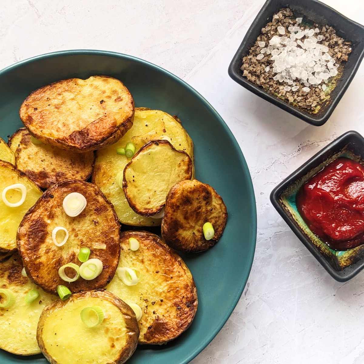 Air fryer sliced potatoes on a blue plate with bowls of seasoning and tomato sauce alongside.