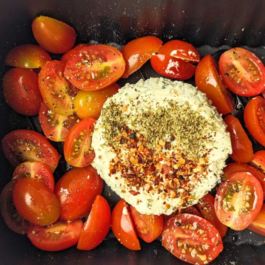 A round of Boursin cheese in an air fryer basket surrounded by halved plum tomatoes.