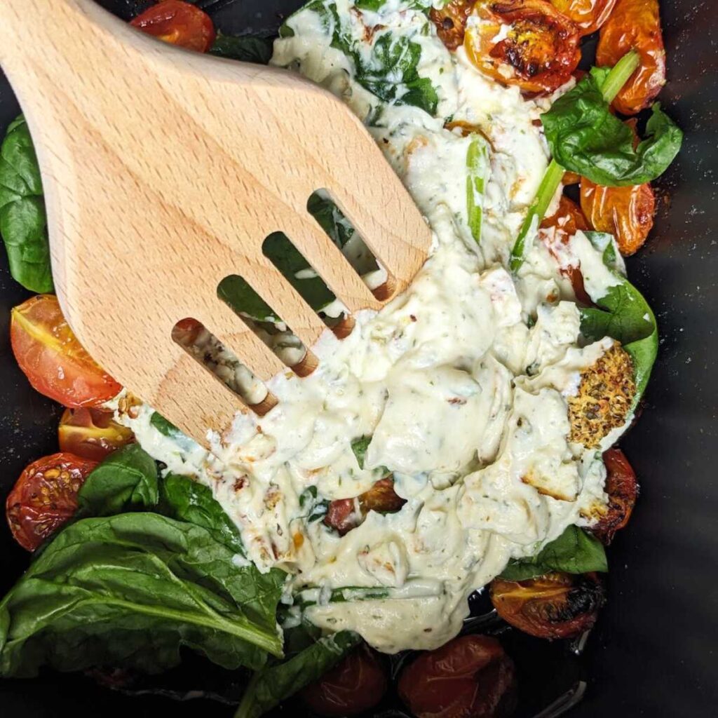 Baked cheese with roasted tomatoes and fresh spinach in an air fryer basket. A wooden spoon is mixing the ingredients together.