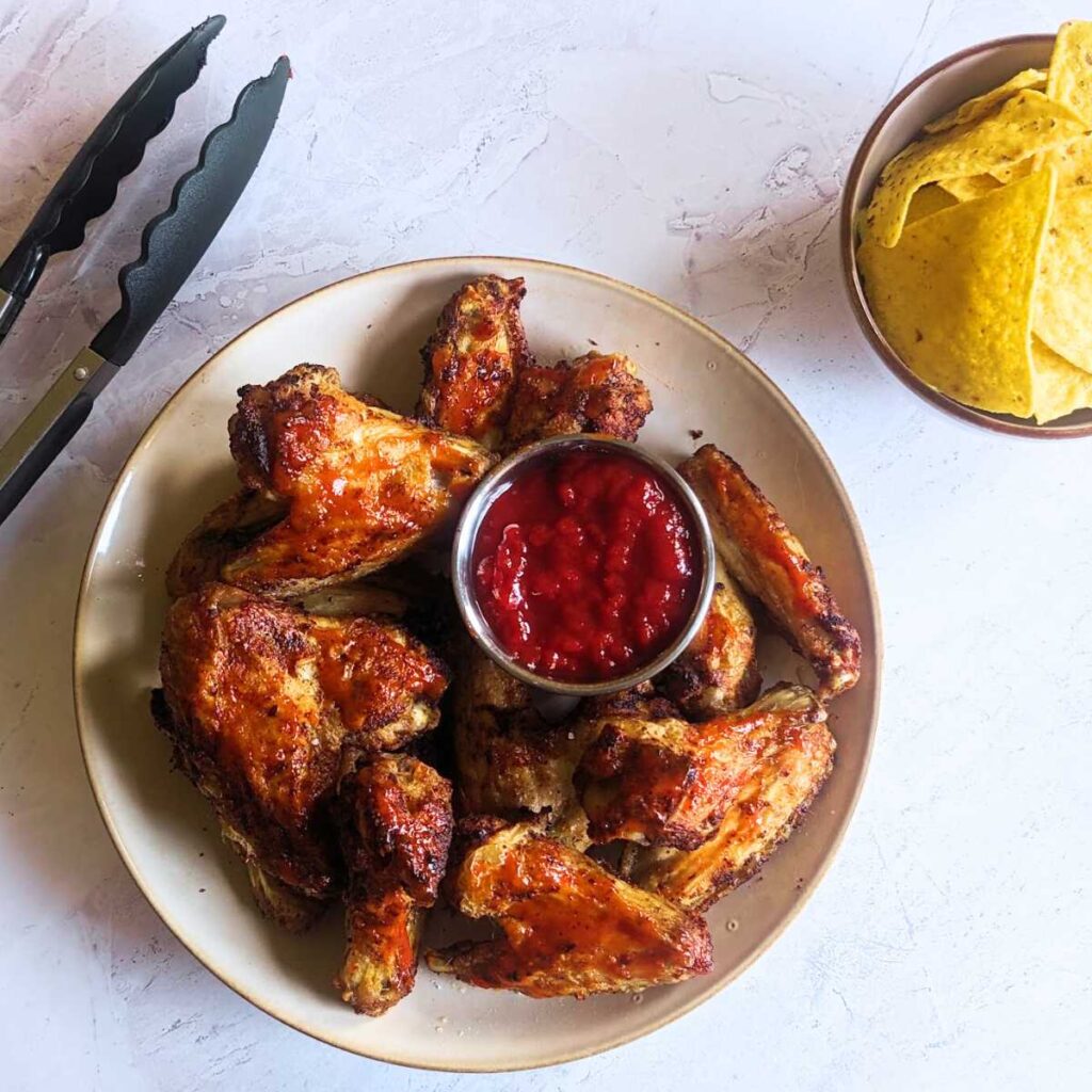 A plate of chicken wings with a tomato salsa. A small bowl of tortilla chips sits at the side.